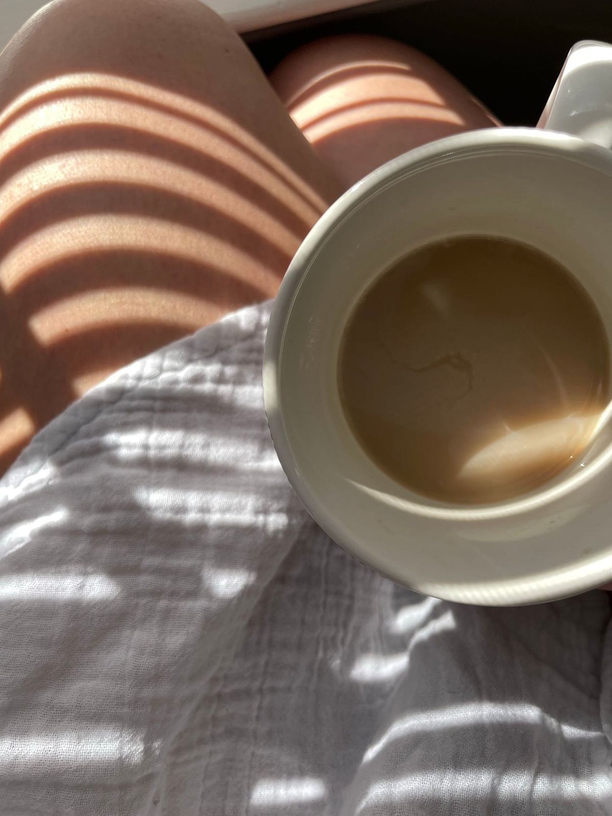 Mok koffie op schoot op een mousseline doek, met strepen ochtendzon door de jaloezie. Blog Rosie's Naturals